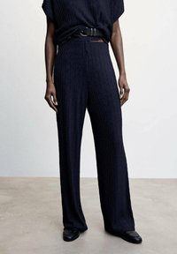 Pantalons larges plissés bleu marine avec une finition texturée et une taille ajustée. Associés à un haut ample et une ceinture noire, complétés par des chaussures noires.