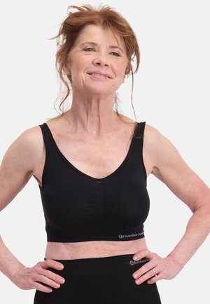 Donna dai capelli rossi con reggiseno sportivo nero e leggings, in piedi con le mani sui fianchi, sorride con sicurezza davanti a uno sfondo semplice.