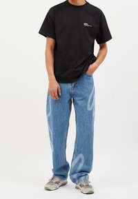 Camiseta de algodón negra con logo blanco en el lado izquierdo del pecho, combinada con unos vaqueros de corte suelto en azul claro que presentan patrones abstractos en blanco.