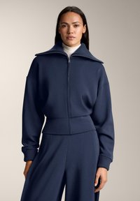 Nainen pukeutuneena laivastonsiniseen vetoketjulliseen takkiin, jossa on leveä kaulus, ja siihen sopiviin leveälahkeisiin housuihin, yksiväristä beigeä taustaa vasten.