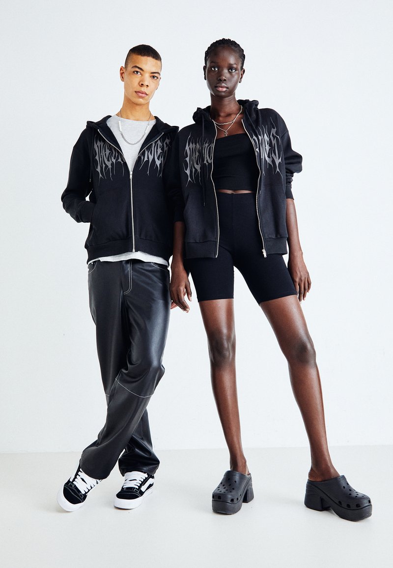 Felpe nere con zip e logo, T-shirt grigia, pantaloni in pelle e scarpe nere. Modello in pantaloni corti e zoccoli con zeppa si trova accanto.