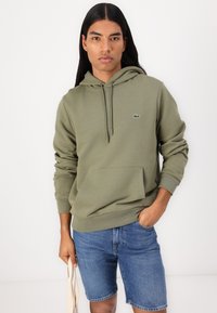 Sweat à capuche vert olive avec cordons et poche avant. Présente des poignets côtelés fins, un logo sur la poitrine, associé à un short en denim.