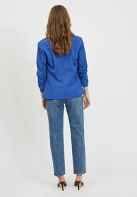 Blå figursydd kavaj med långa ärmar, i kombination med högmidjade ljusblå jeans och svarta klacksandaler. Slätt tyg, skräddarsydd design.