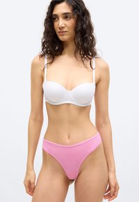 Γυναίκα με σγουρά σκούρα μαλλιά, που φο ράει ένα λευκό σουτιέν χωρίς ράντες και ροζ εσώρουχο με ψηλό κόψιμο, στέκεται μπροστά σε μια απλή ανοιχτόχρωμη επιφάνεια.