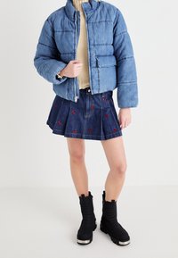 Person wearing a blue puffer jacket, beige sweater, pleated denim skirt with red butterfly embroidery, and black chunky boots standing on white floor.