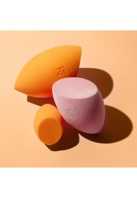 Trois éponges de maquillage orange et rose avec des bords plats et arrondis sur un fond pêche projetant des ombres.