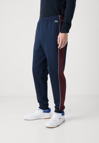 Marinblå joggingbyxor med en bordeauxröd sidopanel och vit kantning, gjorda av mjukt tyg. Bärs med vita sneakers med blåa detaljer.