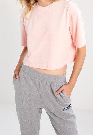 Mujer con un top corto de manga corta en rosa pálido y pantalones deportivos grises con las manos en los bolsillos, de pie frente a un fondo claro liso.