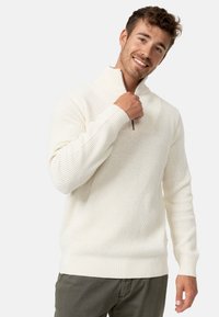 Suéter de punto blanco con cremallera de quartzo y patrón texturizado. Presenta puños y dobladillo acanalados. Llevado con un par de pantalones de color neutro.