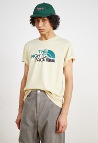 Beige bomulds T-shirt med rullede ærmer, der har et teal og sort "The North Face" logo, parret med grå kordfløjl bukser. Grøn kasket på hovedet.