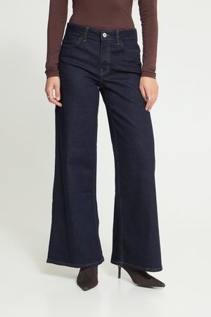 Femme portant un jean large bleu foncé, un haut à manches longues marron et des bottines à talons pointues, debout devant un fond uni.