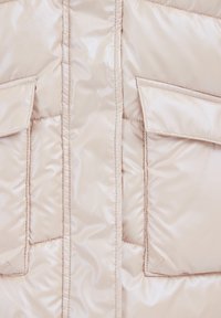 Beige puffjacka med en glansig finish, med två lapplommer och sydda detaljer längs sömmarna. Mjuk, slät textur.