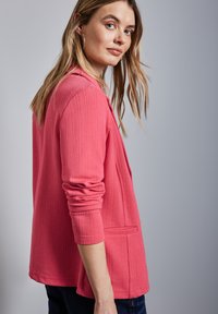 Teksturowana, lekka, różowa marynarka z otwartym przodem, długimi rękawami i kieszeniami po bokach. Modelka zaprezentowana z boku.