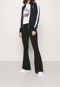 Sort lynlåsjakke med hvide striber, hvid grafisk t-shirt og sorte flagrende bukser. Modelen bærer hvide sneakers.