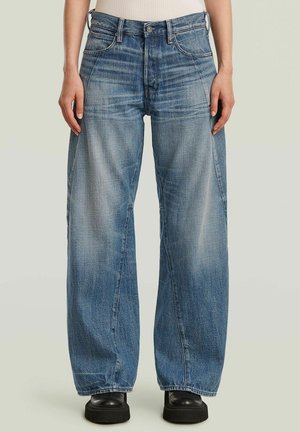 Personne portant un jean bleu délavé à jambes larges et des bottes noires à semelle épaisse, debout face à un fond uni.