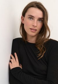 Zwarte langemouwen shirt met een gladde textuur, gedragen door een persoon die tegen een witte muur leunt. Het haar is middellang en bruin.