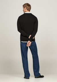 Schwarzer gestrickter Pullover mit langen Ärmeln, getragen über einem weißen Hemd, kombiniert mit blauen Jeans. Rückansicht, gerade geschnitten mit minimalen Details.