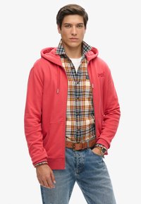 Koraalrode hoodie met voorkruiszakken en een verstelbare capuchon over een geruit flanellen overhemd. Draagt een lichte spijkerbroek en een bruine riem.