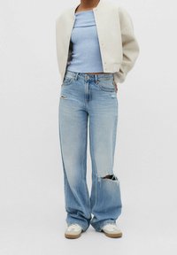 Hellblau geripptes Croptop unter einer cremefarbenen Bomberjacke, kombiniert mit weit geschnittenen, stark ausgewaschenen Jeans und beigen Sneakern.