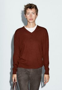 Maglione a V, dal taglio morbido, in pregiato tessuto di lana marrone abbinato a pantaloni grigi slim. Il modello porta una borsa marrone.