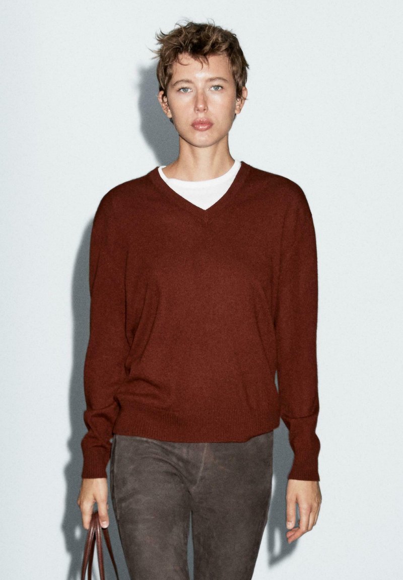 Maglione a V, dal taglio morbido, in pregiato tessuto di lana marrone abbinato a pantaloni grigi slim. Il modello porta una borsa marrone.