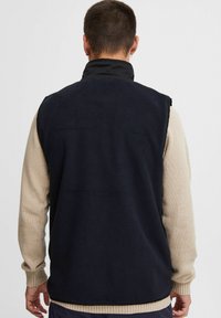 Chaleco de forro polar azul marino oscuro con un cuello alto y sin mangas, que presenta una capa base de manga larga beige y puños de punto texturizado.