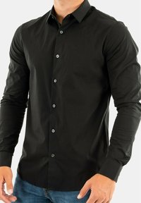 Mężczyzna w dopasowanej czarnej koszuli z długimi rękawami i niebieskich dżinsach na jednolitym tle.