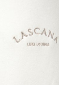 Weißer Stoff mit beige gesticktem Text, der "LASCANA LUXE LOUNGE" liest, und eine glatte, weiche Textur zeigt.