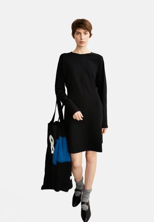 Zwarte lange mouw jurk met een ronde halslijn, een gladde textuur, gecombineerd met grijs gestreepte sokken en zwarte schoenen, terwijl ze een zwarte draagtas met blauwe accenten vasthoudt.
