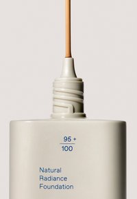 Biała prostokątna butelka z końcówką aplikatora. Niebieski tekst pokazuje "95 + 100" oraz "Natural Radiance Foundation". Gładkie, matowe wykończenie.