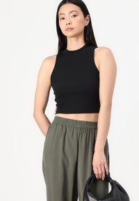 Top court sans manches noir avec un col montant associé à un pantalon ample vert olive. Le mannequin tient un sac à main noir avec une anse arrondie.