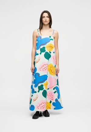 Nő áll, hosszú fehér ruhát visel, amely nagy kék, rózsaszín, sárga és zöld virágmintákkal díszített, fekete cipőben, egyszínű háttér előtt.