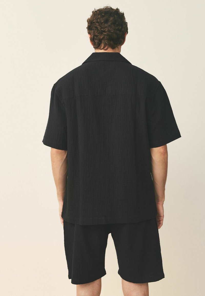 Mand med krøllet hår iført løs sort kortærmet skjorte og matchende shorts, stående mod en ensfarvet lys baggrund, med ryggen mod beskueren.