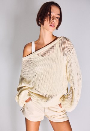 Mladý človek s krátkymi vlasmi, ktorý má na sebe voľný krémový sveter s odhalenými ramenami cez biely tielkový top a krémové šortky, ruky v kapse.