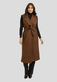 Gilet long en mélange de laine brun avec de grands revers, taille ceinturée et deux poches avant. Porté sur un pull à col roulé noir et un pantalon noir.