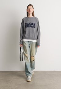 Suéter cinza de malha com texto azul escuro, emparelhado com jeans claros desgastados. Sapatos prateados e uma bolsa metálica completam o visual.