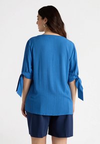 Blusa blu in crepe con tessuto testurizzato, scollatura rotonda e maniche legate. Abbinata a pantaloni corti blu navy, che mostrano una vestibilità ampia e rilassata.