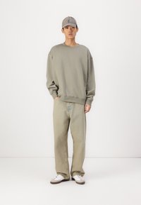 Sweatshirt vert olive surdimensionné, jean large beige clair, et casquette grise. Baskets blanches avec accents marron visibles aux pieds.