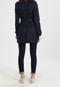 Marinblå trenchcoat med bälte i midjan, knappdetaljer och hög krage, kombinerad med mörka jeans och ljusa skor, med uppvikta muddar.