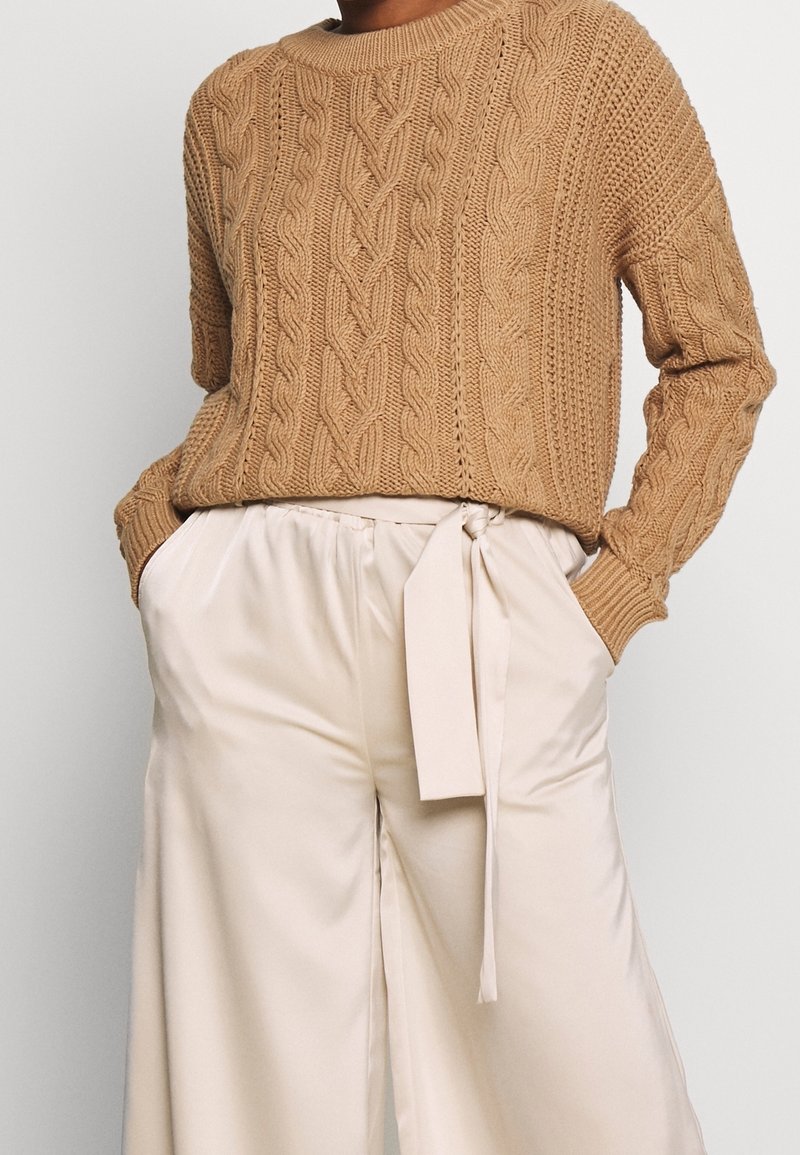 Beige cable-knit sweater with a round neck and ribbed cuffs, paired with pale satin wide-leg trousers featuring a tied waist.