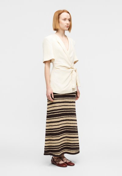 Μπεζ μπλούζα με ζώνη και κοντά μανίκια, συνδυασμένη με καφέ ριγέ πλεκτή μάξι φούστα. Καφέ σανδάλια με λουράκια ολοκληρώνουν την εμφάνιση.
