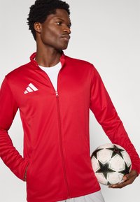 Κόκκινο αθλητικό μπουφάν με φερμουάρ μπροστά, μακριά μανίκια και πλαϊνές τσέπες. Διαθέτει λευκό λογότυπο Adidas και ελαφρύ, λείο ύφασμα. Κρατά μια ποδοσφαιρική μπάλα.