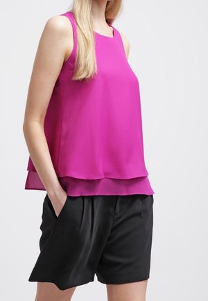 Femme portant un haut sans manches rose magenta à plusieurs couches avec un short noir plissé, debout avec une main dans la poche, sur un fond uni.