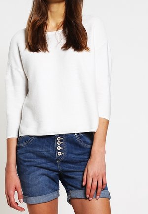 Femme portant un haut blanc côtelé à manches longues et un short en jean bleu taille haute avec cinq boutons visibles.