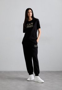Zwarte katoenen t-shirt met gouden logo, gecombineerd met zwarte joggers met een bijpassend logo, en witte sneakers. Gladde textuur, relaxte pasvorm.