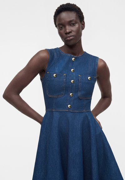 Γυναίκα που φοράει μπλε φόρεμα από τζιν χωρίς μανίκια με χρυσά κουμπιά και τσέπες στο στήθος, στέκεται με τα χέρια πίσω από την πλάτη σε απλό φόντο.