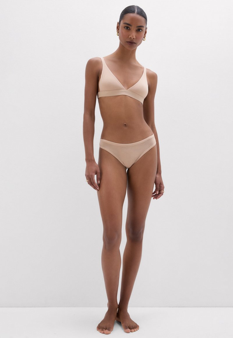Mujer de pie frente a un fondo blanco liso, lleva un bralette color nude y ropa interior a juego, mirando al frente con los brazos relajados a los lados.