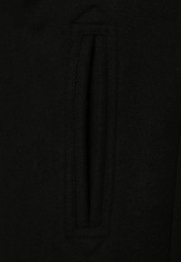 Abrigo de lana negro con un bolsillo vertical con parte superior redondeada, tejido texturizado y costuras limpias sin herrajes visibles.