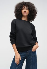 Γυναίκα με σγουρά μαλλιά που φοράει απλό μαύρο φούτερ και μπλε τζιν, στέκεται με το ένα χέρι στην τσέπη μπροστά σε απλό φόντο.