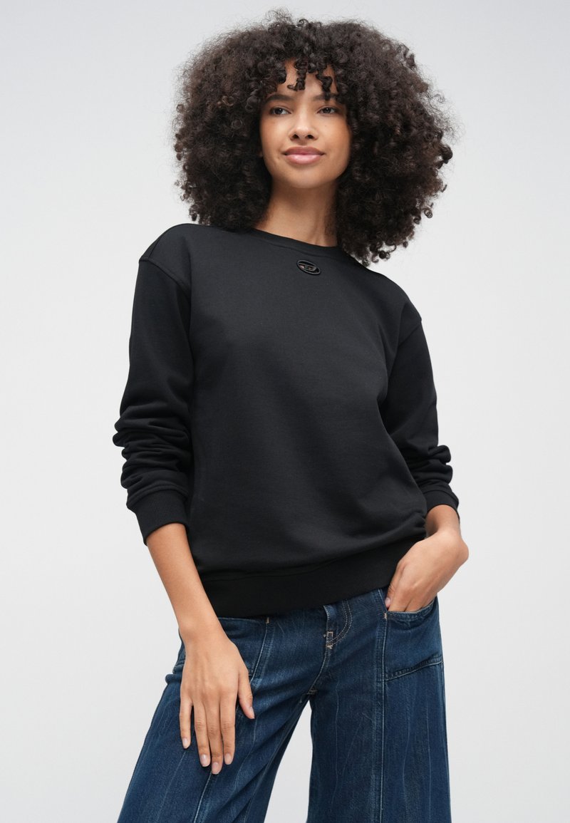 Donna con capelli ricci che indossa una felpa nera semplice e jeans blu, in piedi con una mano in tasca contro uno sfondo semplice.